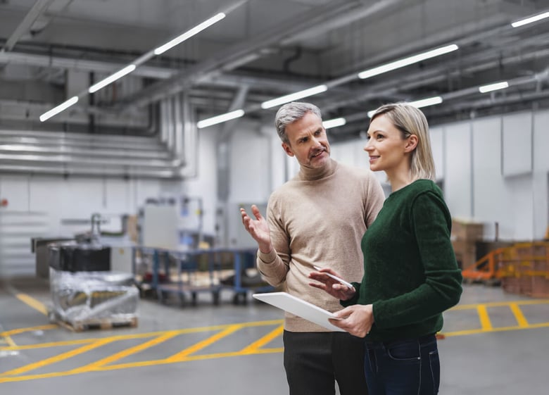 Two Syntegon employees talking in an industrial workspace, one holding documents.