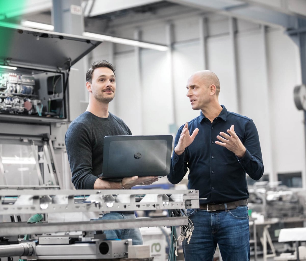 Zwei Ingenieure im Gespräch vor einer Industrieanlage, einer hält einen Laptop.