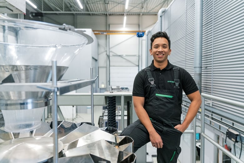 Syntegon technician smiling beside packaging machinery in a modern production facility.